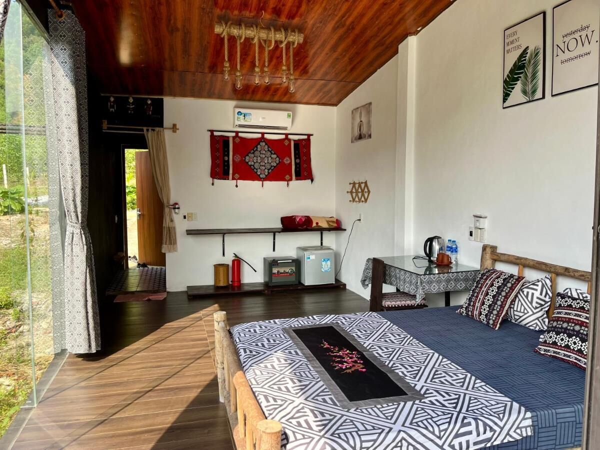 Quarto com cama, uma pequena mesa e uma cadeira. Há uma janela com cortinas, um ar condicionado fixado na parede e um objeto decorativo vermelho.