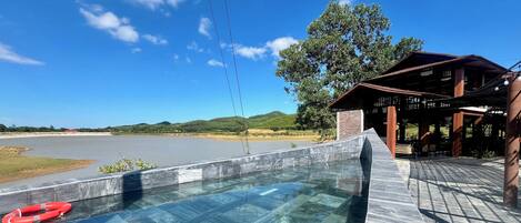 Vista despejada de una piscina con un flotador salvavidas, una terraza de madera y un edificio con techo que da a un cuerpo de agua y colinas.