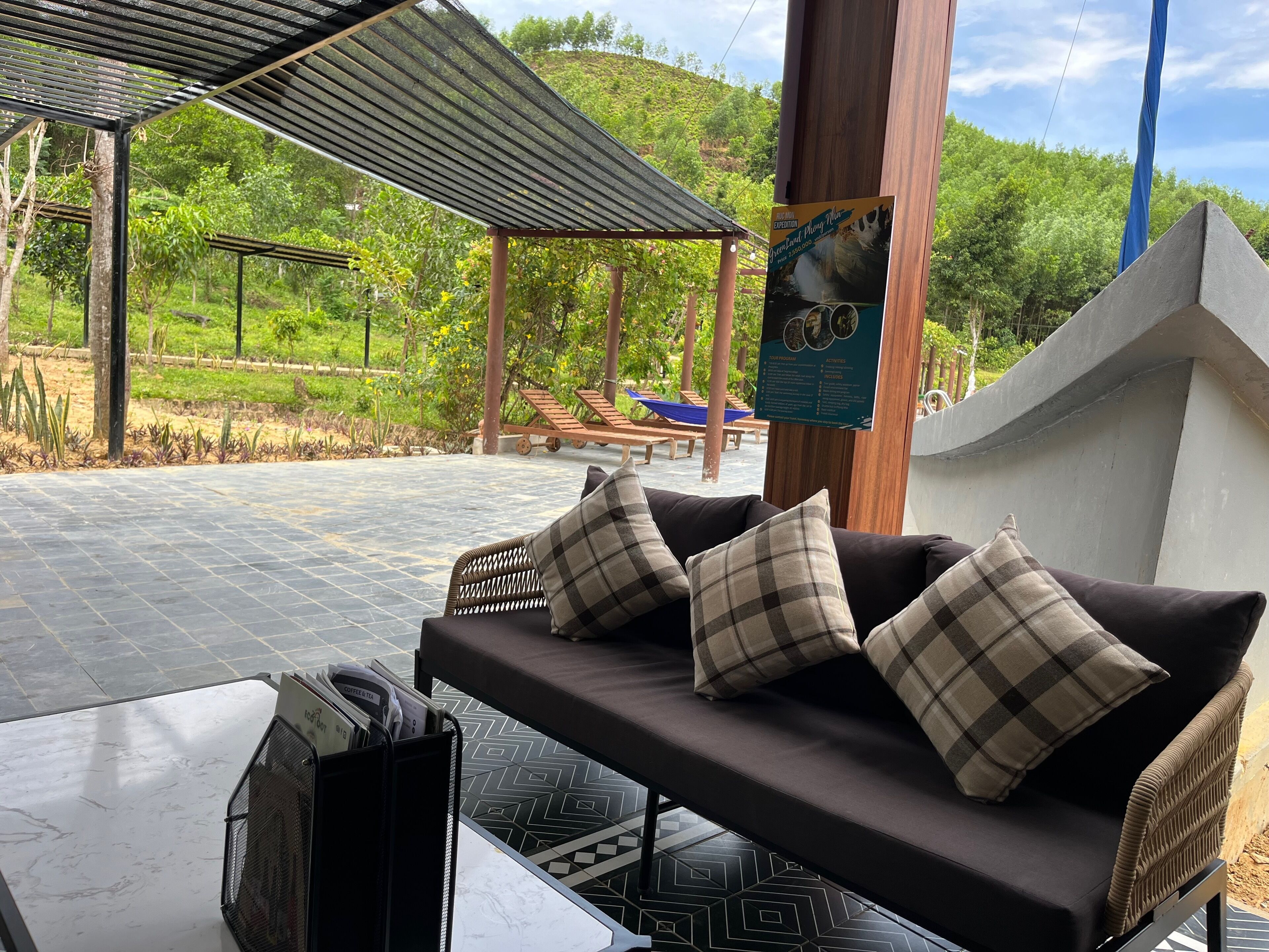 An outdoor seating area with a sofa and cushions, a pergola, and a view of a lush garden and hills.