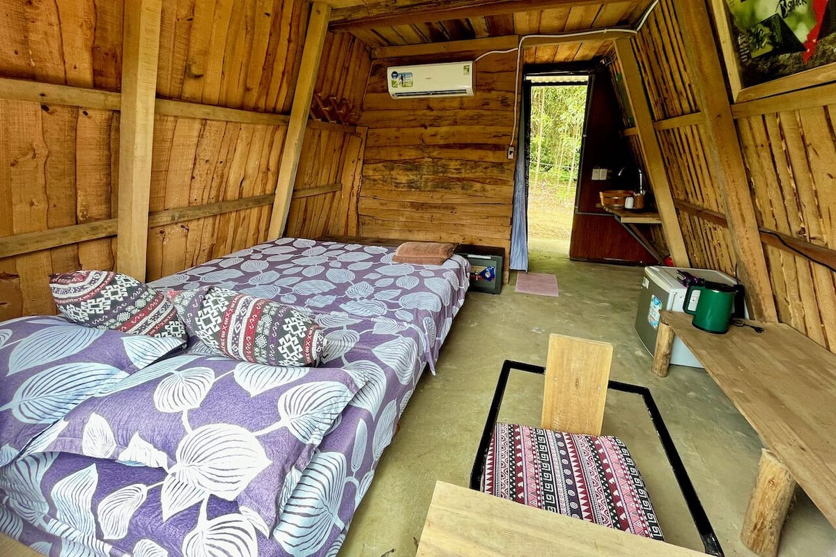 A wooden cabin interior with a bed, a small table, and a bench.