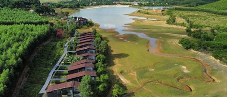 An aerial view of a resort with a long row of buildings along a river, surrounded by lush greenery and hills.