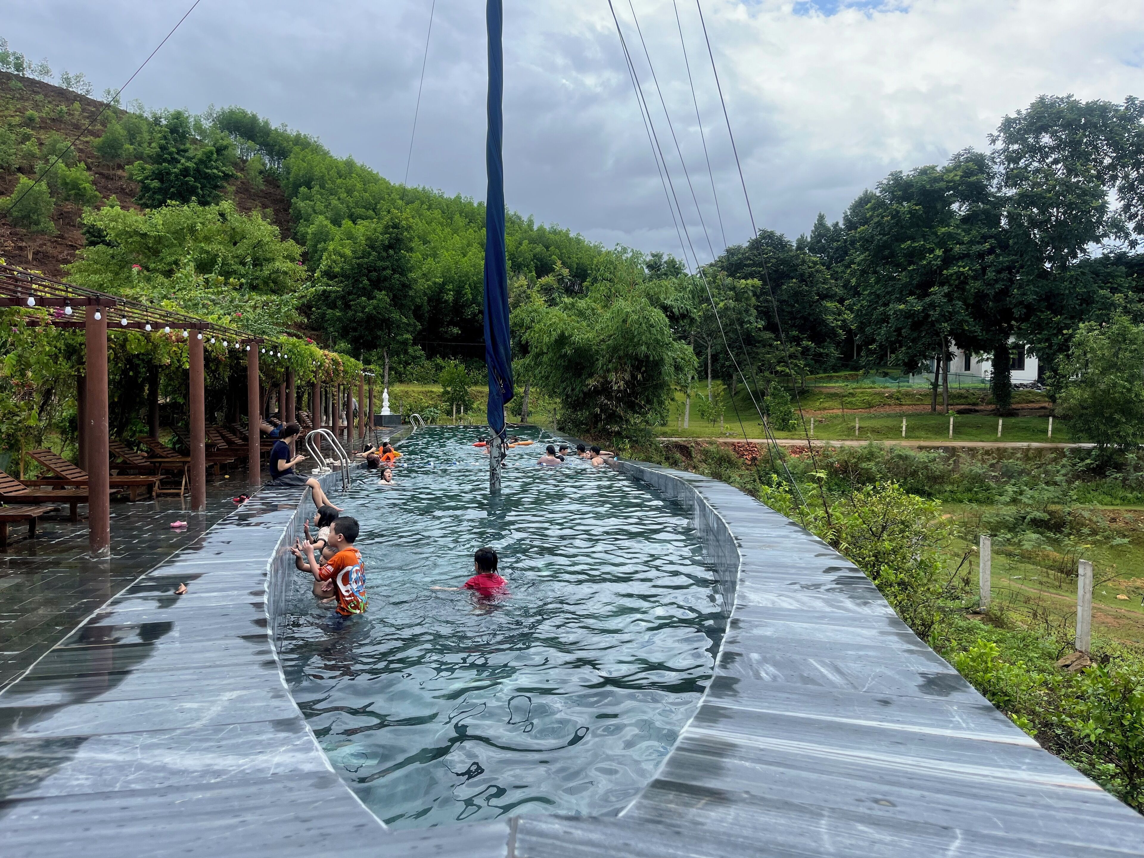 Une piscine où l’on voit des personnes, entourée de verdure et surmontée d’une pergola illuminée.