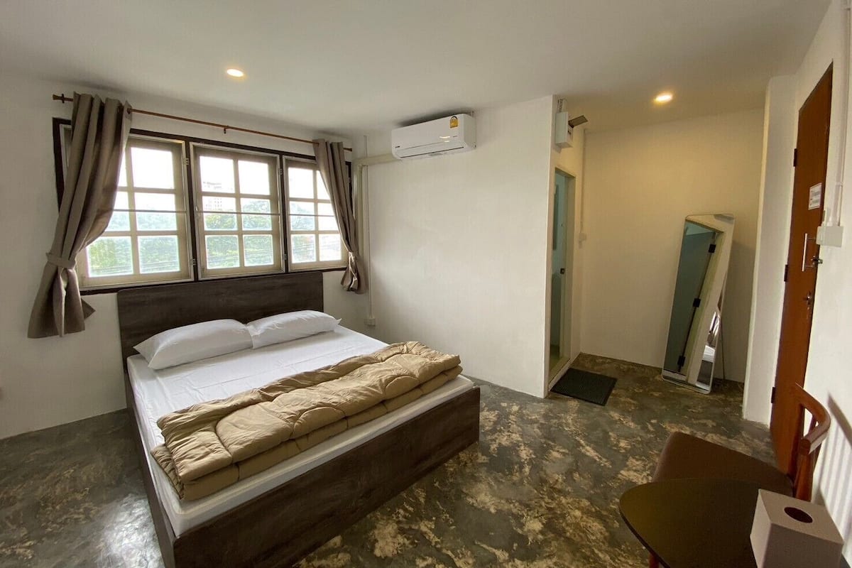 A bedroom with a bed, a window with curtains, an air conditioning unit, and a small table with a tissue box.