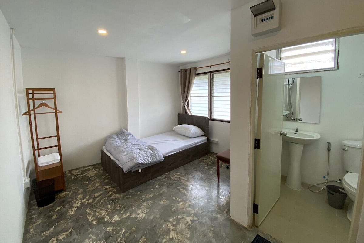A small, single-bed room with a wooden ladder, a window with curtains, a bathroom with a sink and toilet, and a concrete floor.