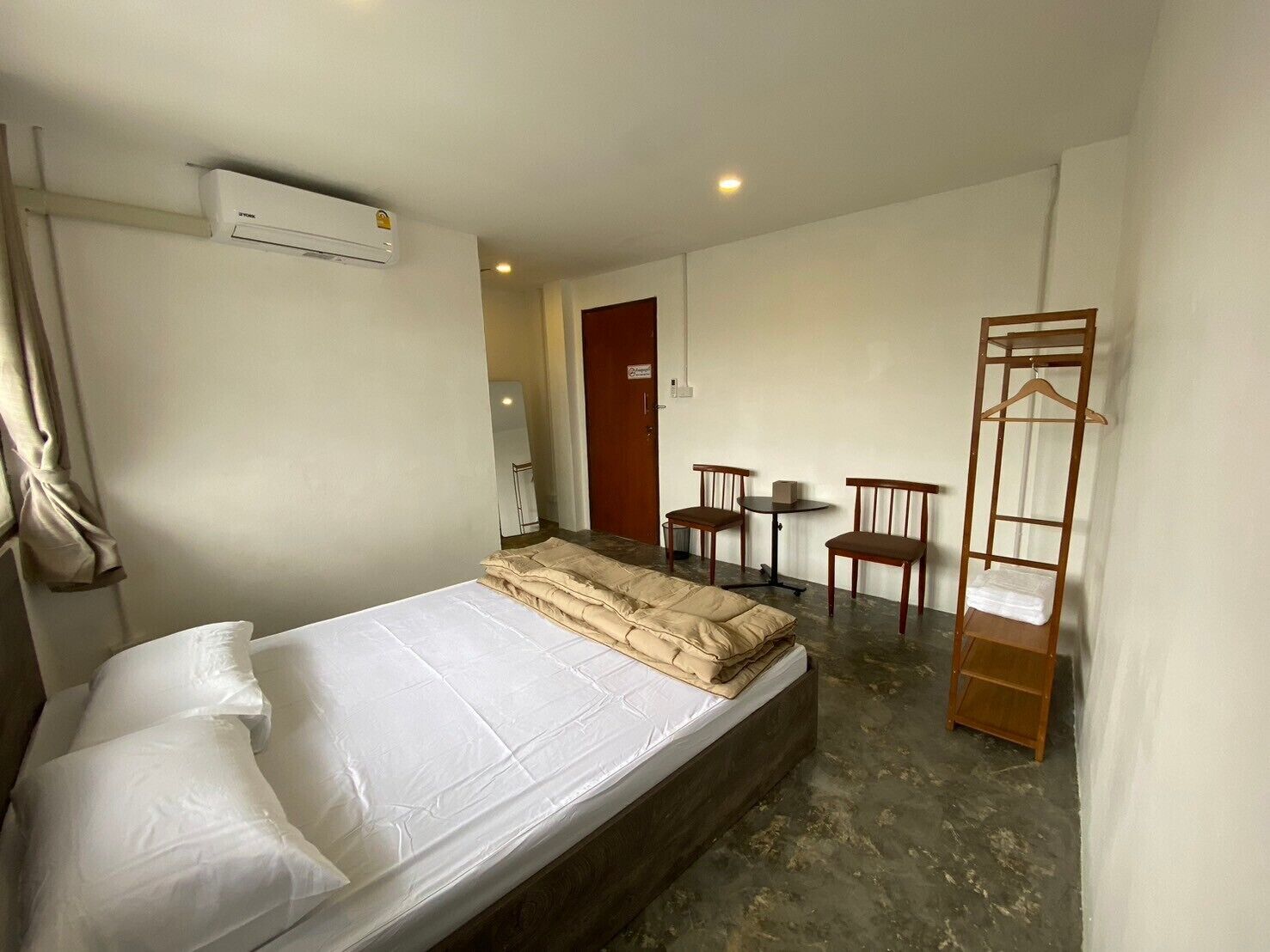 A single bed with a white mattress and pillows, a wooden bedside table, a small wooden chair, a wooden chair at a small round table, and a wooden wardrobe with a hanging shelf.