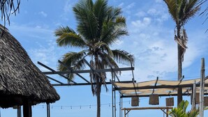 A beachside resort with a thatched-roof structure, wooden deck seating, and palm trees.