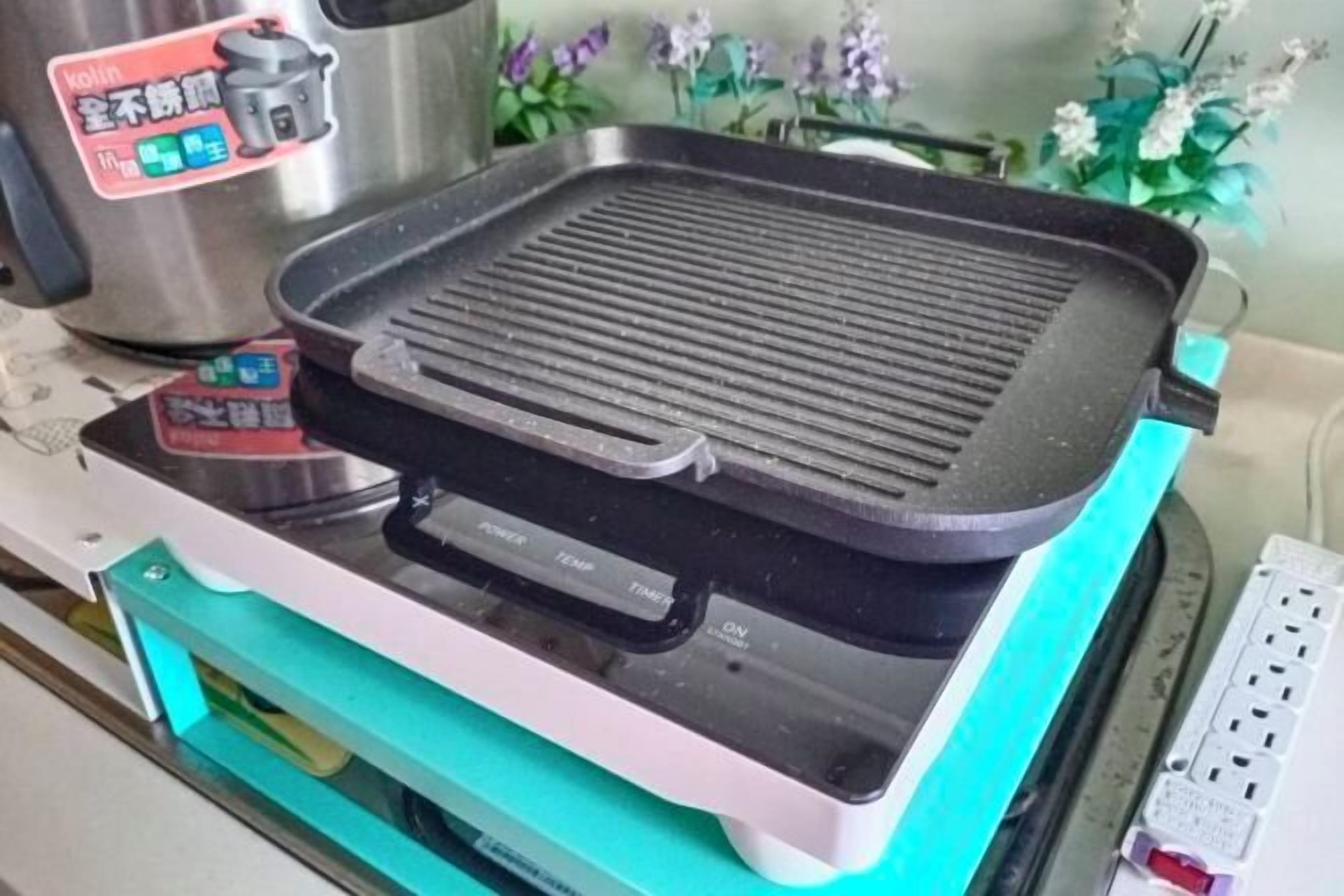 Una cucina con una piastra grill su un piano cottura a induzione, una pentola in acciaio inox e una piccola scatola rossa con caratteri cinesi.