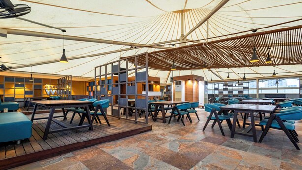 A spacious indoor area with tables and chairs, a wooden floor, and a high ceiling with a unique design. - Treebo Blue Moon (Gandhinagar)