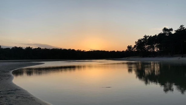 Um lago calmo ao pôr do sol, com uma praia de areia e uma floresta ao fundo.