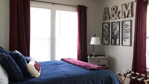 A bedroom with a bed, red curtains, a nightstand with a lamp, and wall art. - 7 Minute Walk to Kyle Field - Charming Cottage (College Station)
