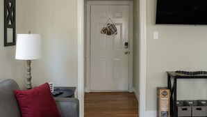 A living room with a grey sofa, a red pillow, a lamp, a television, and a door with a football-themed decoration. - 7 Minute Walk to Kyle Field - Charming Cottage (College Station)