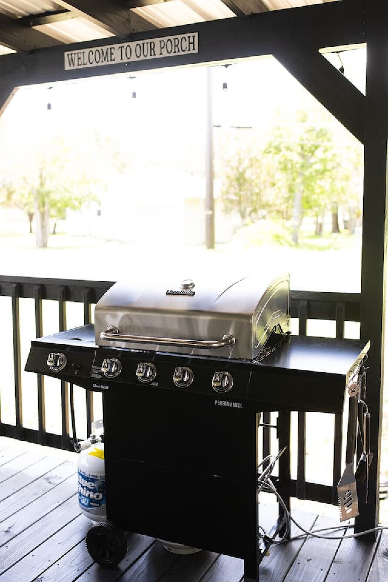 Ein Grill mit einem 'Welcome to Our Porch' Schild darüber, platziert auf einer Holzterrasse, in der Nähe steht eine Wasserflasche.