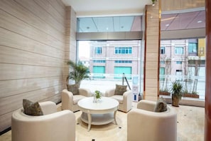 Un hall d’entrée moderne avec une table ronde et quatre canapés, de grandes fenêtres offrant une vue sur les bâtiments et des plantes en pot.