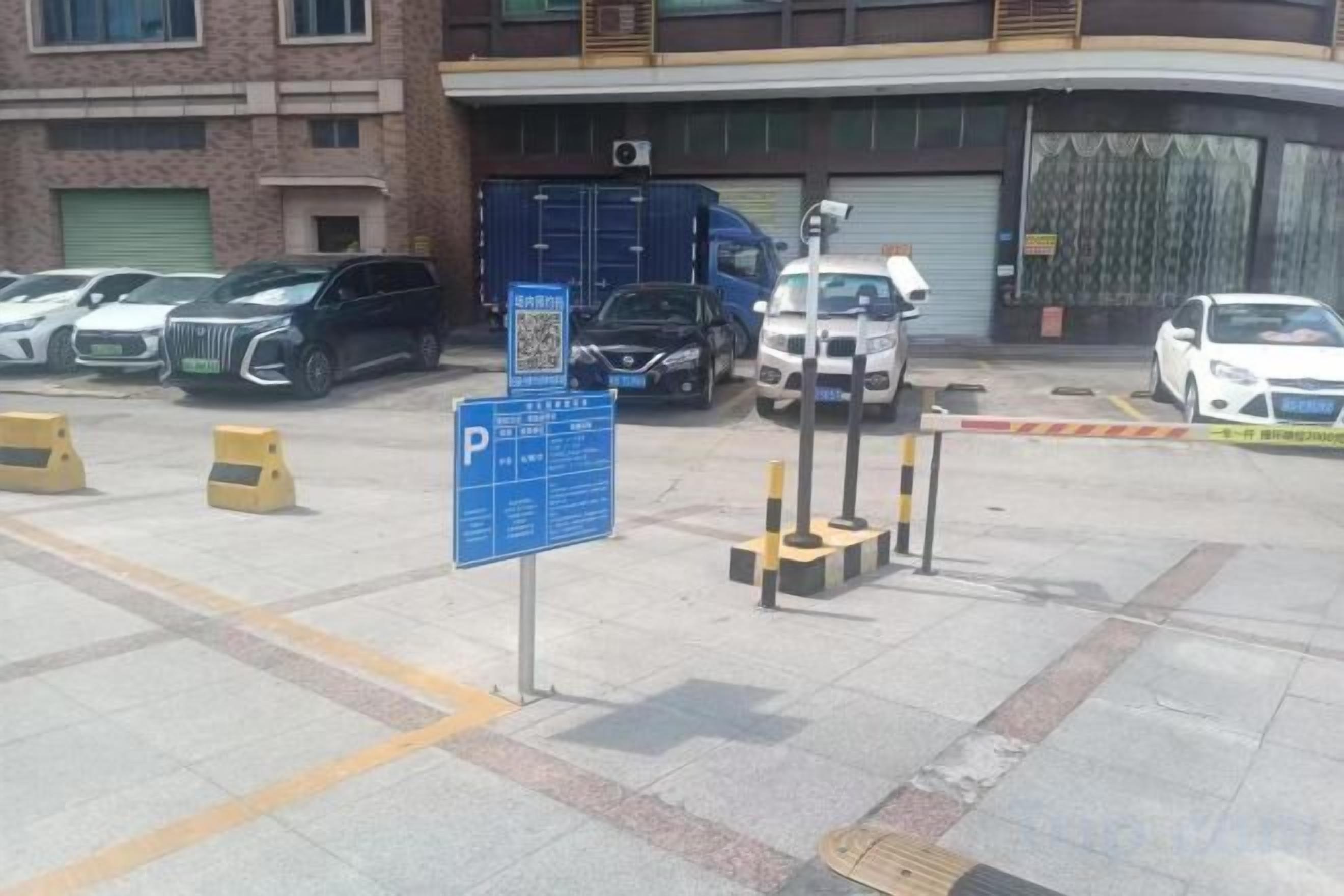 A parking lot with multiple cars, a blue parking sign, and a building with a garage door in the background.