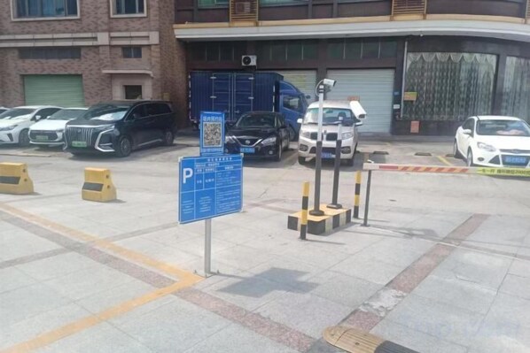 A parking lot with multiple cars, a blue parking sign, and a building with a garage door in the background. - Weisidun Hotel (Dongguan)