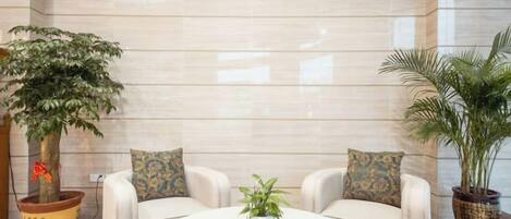Een ontvangsthal met een marmeren wand, twee banken met kussens, een ronde salontafel en potplanten.
