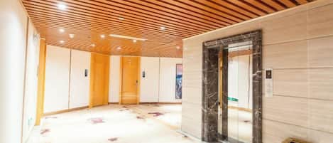 A hotel corridor with wooden panel ceilings, marble walls, and a central elevator.