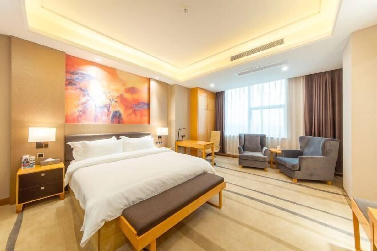 Ein Hotelzimmer mit einem Bett, einer Bank, einem Schreibtisch, zwei Sesseln, einem großen abstrakten Gemälde und einem Fenster mit Vorhängen.