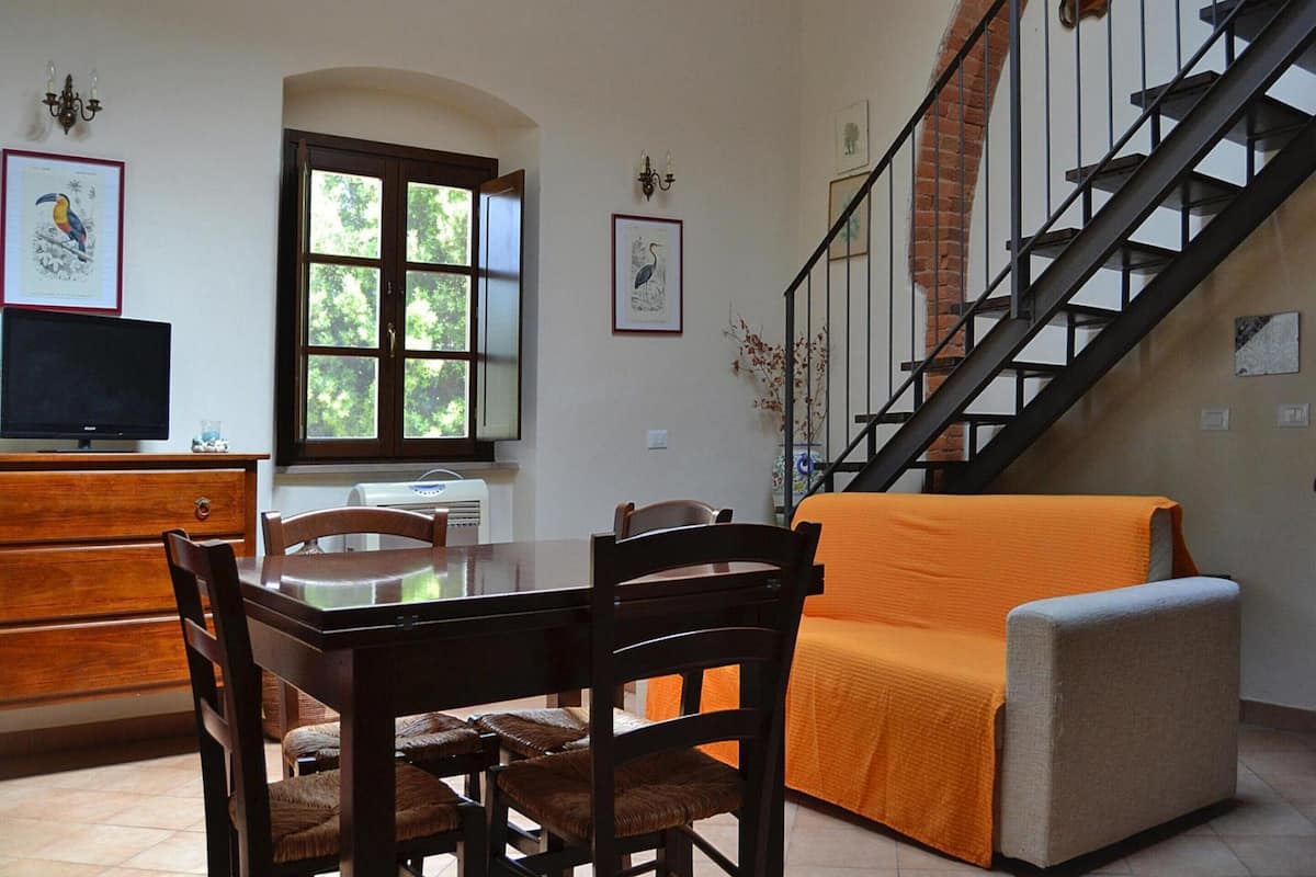 Ein Essbereich mit Holztisch und -stühlen, einer Couch mit orangefarbener Decke, einem Fernseher auf einem Holzkasten und einer Treppe an einer Backsteinwand.