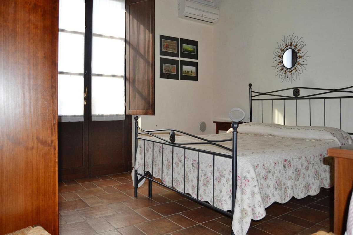 Ein Schlafzimmer mit Holzboden, einem Bett mit Blumenmuster-Kopfbekleidung, einem Wandspiegel und einem Fenster mit Vorhang.