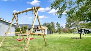 Ein hölzernes Schaukelgerüst in einem grünen Garten mit einem Haus und einem Schuppen im Hintergrund.