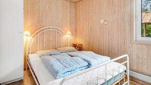 Ein Schlafzimmer mit Holzbalkendecke und -wänden, einem weißen Bett mit geschwungenem Kopfteil, zwei Nachttischlampen und einem Fenster mit Blick auf Grünflächen.
