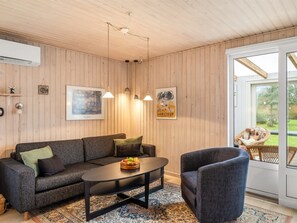 Ein gemütliches Wohnzimmer mit einer Couch, einem Couchtisch und einem Sessel. An den Wänden sind Lampen angebracht und es gibt ein großes Fenster.