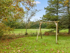 Eine Holzschaukel auf einer grünen Wiese, umgeben von Bäumen und unter blauem Himmel.