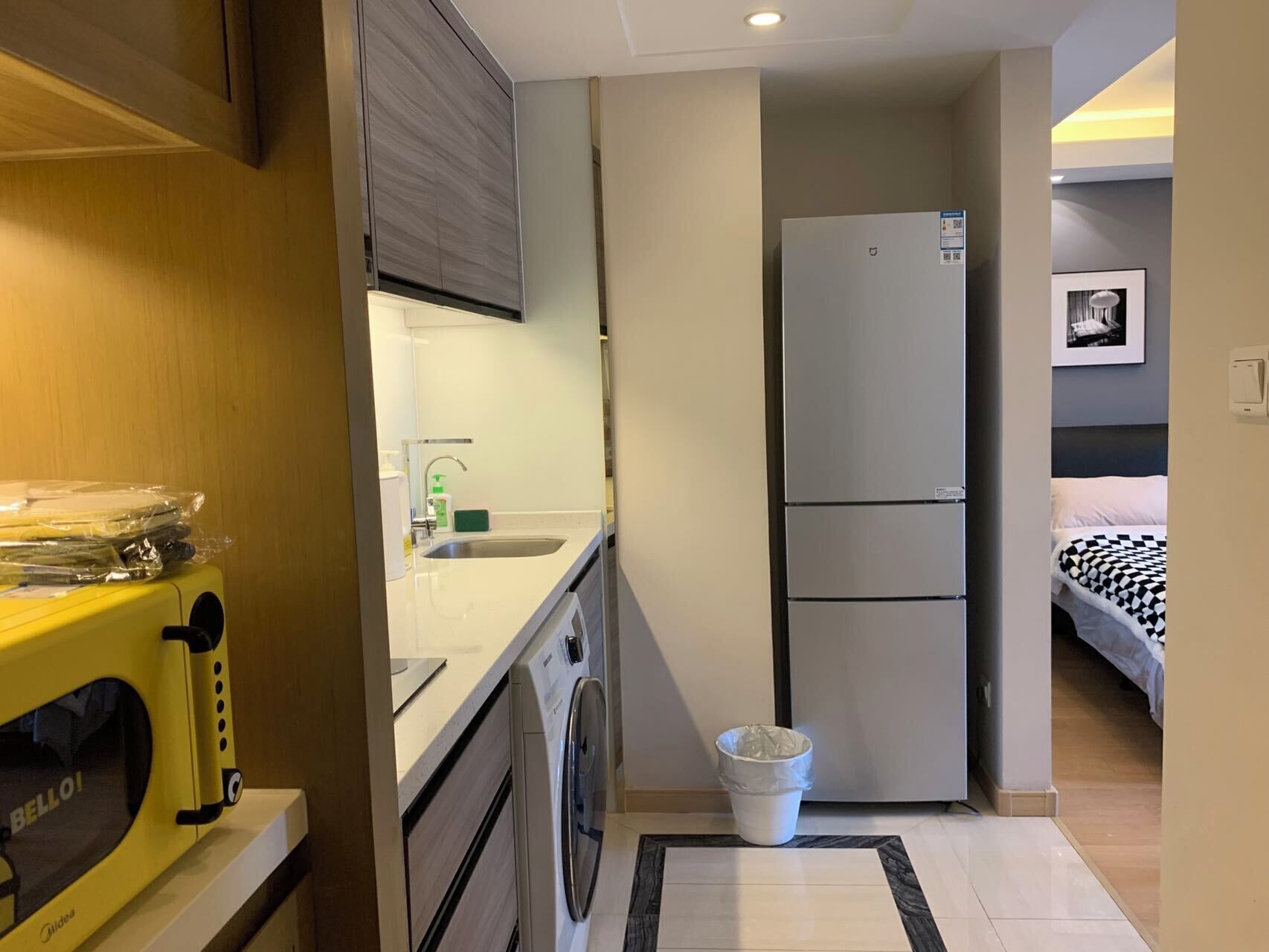 A compact hotel room with a kitchenette, a washing machine, a refrigerator, and a bed with a checkered bedspread.