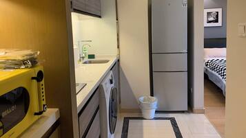 A compact hotel room with a kitchenette, a washing machine, a refrigerator, and a bed with a checkered bedspread.