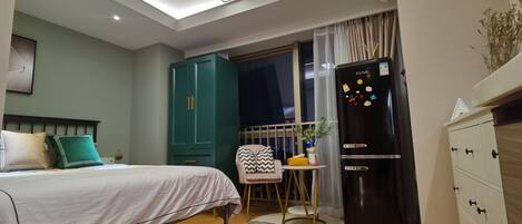 A modern bedroom with a bed, a small table, a chair, a refrigerator, and a wardrobe.