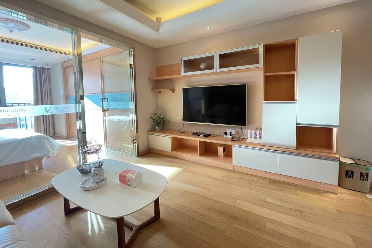 Une chambre d’hôtel moderne dotée d’un grand téléviseur à écran plat, d’étagères en bois, d’une porte en verre donnant sur un balcon et d’une table basse blanche.