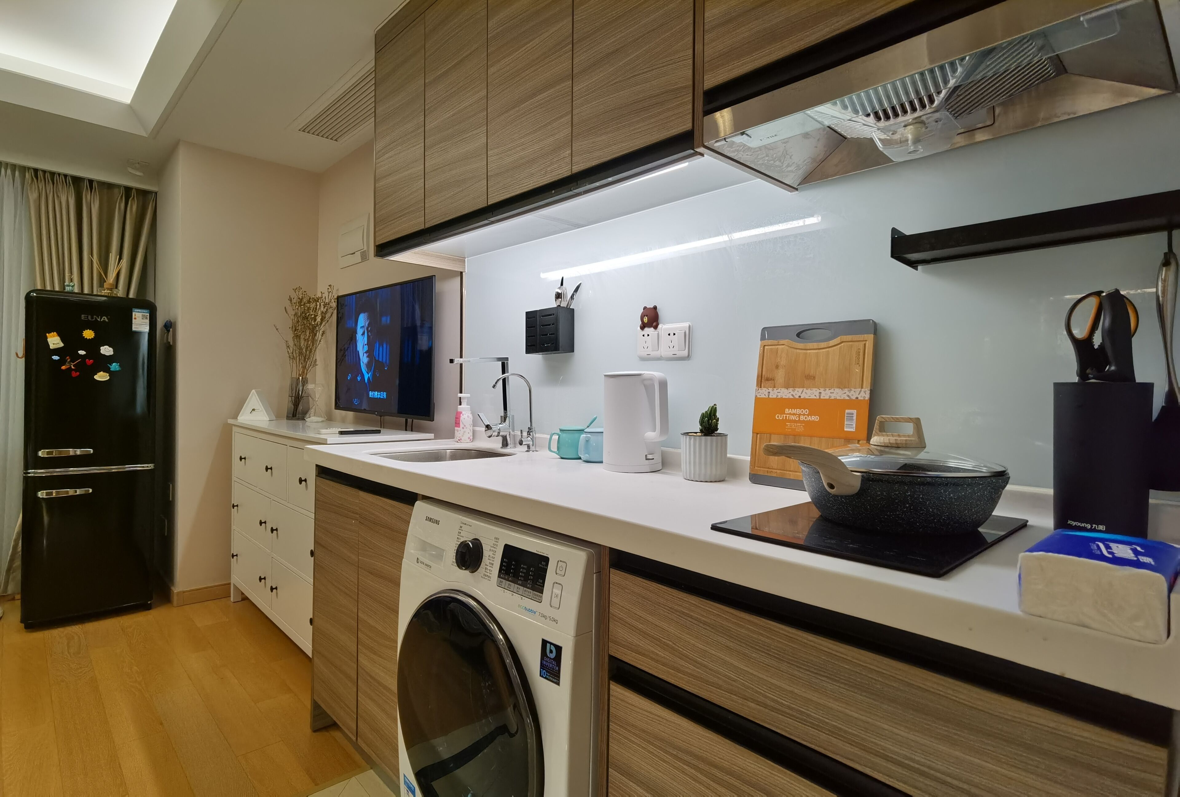 A modern kitchen with a washing machine, a refrigerator, and a flat-screen TV.