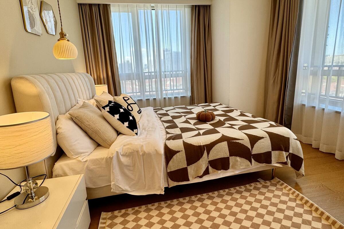 Une chambre d’hôtel avec un lit, une table de chevet avec une lampe, un tapis à carreaux et une vue sur la ville grâce à de grandes fenêtres.
