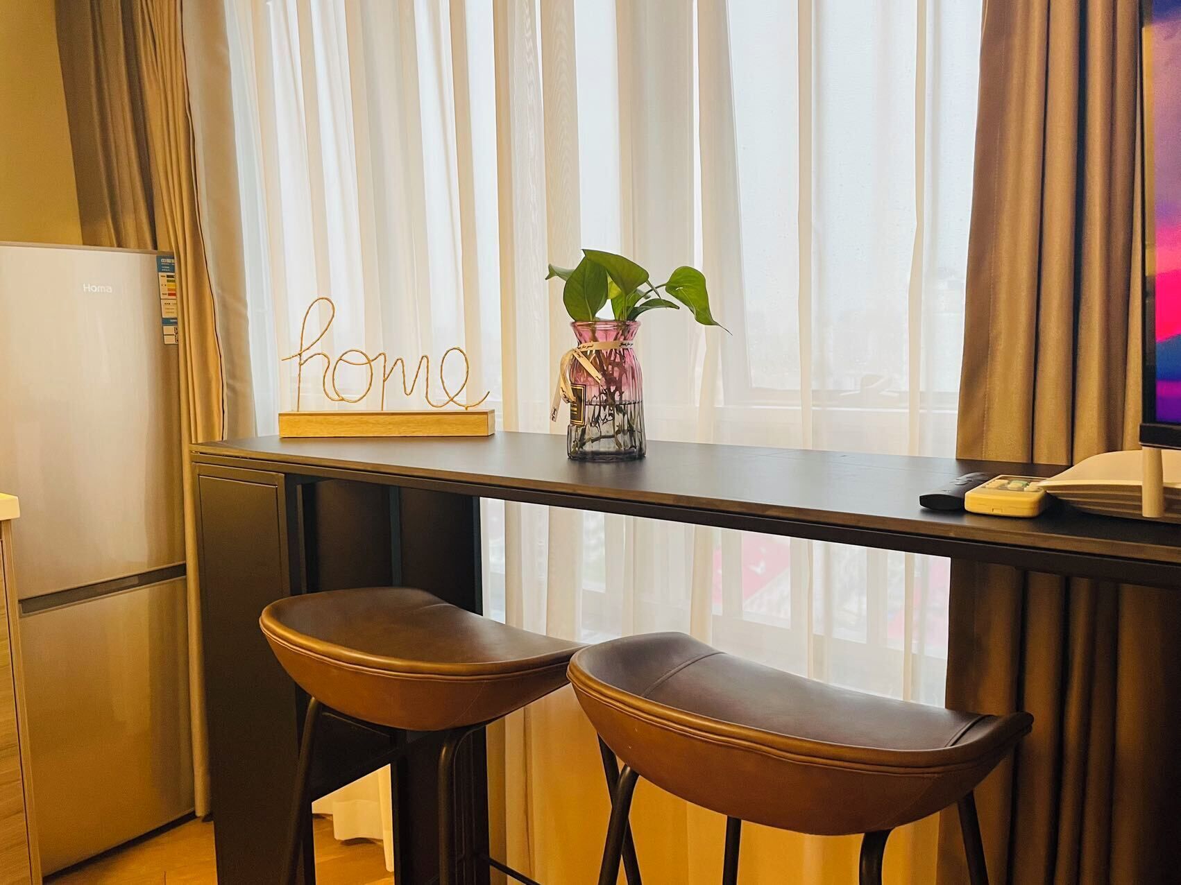 A kitchen with a wooden bar counter, two leather bar stools, a potted plant, and a sign that reads "home".