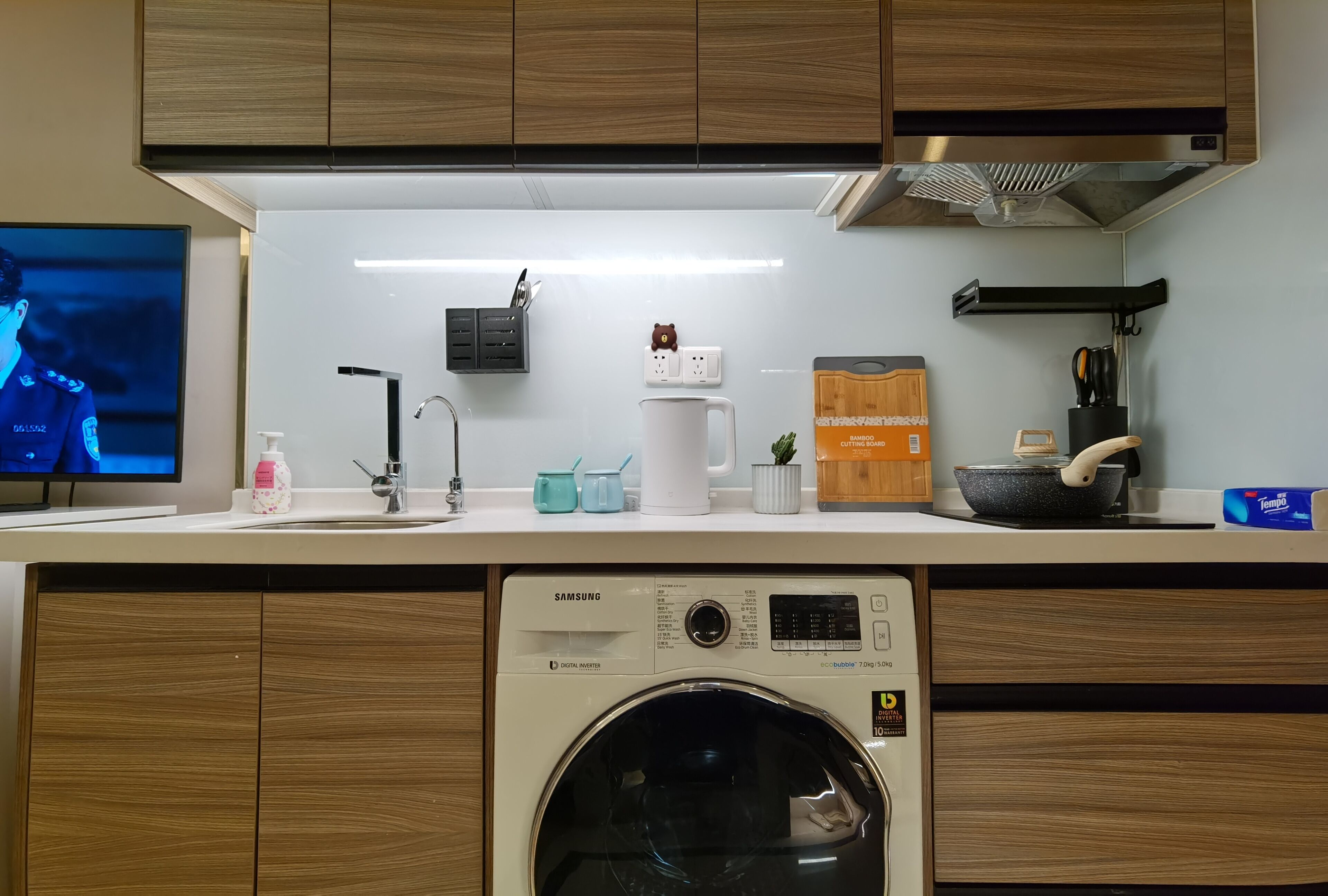 A modern kitchen with wooden cabinets, a built-in washing machine, a sink with a faucet, a television, and various kitchen items on the countertop.