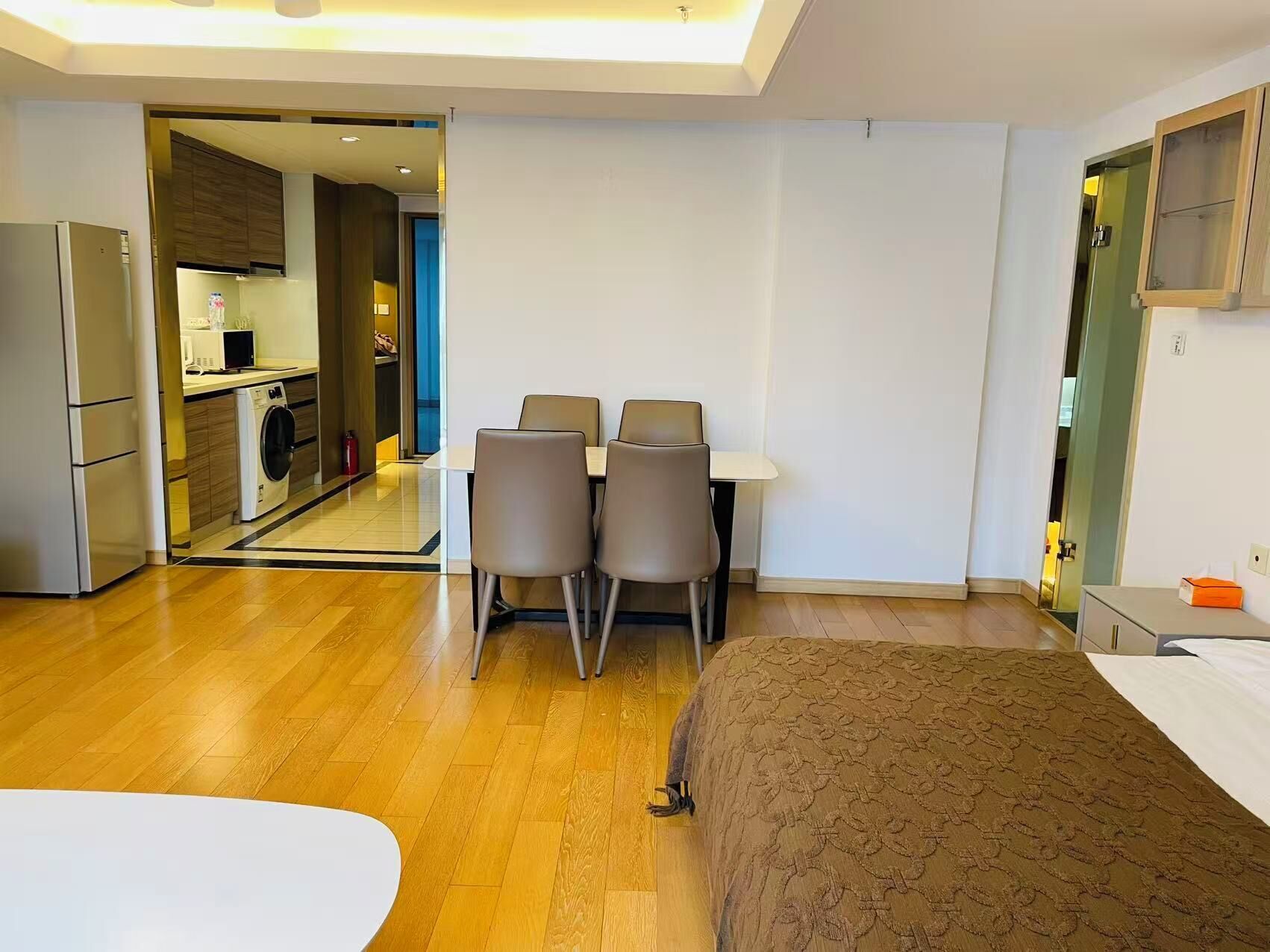 A modern hotel room with a bed, a dining table with chairs, a kitchen area, and a bathroom.