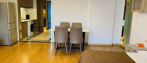 A modern hotel room with a bed, a dining table with chairs, a kitchen area, and a bathroom.