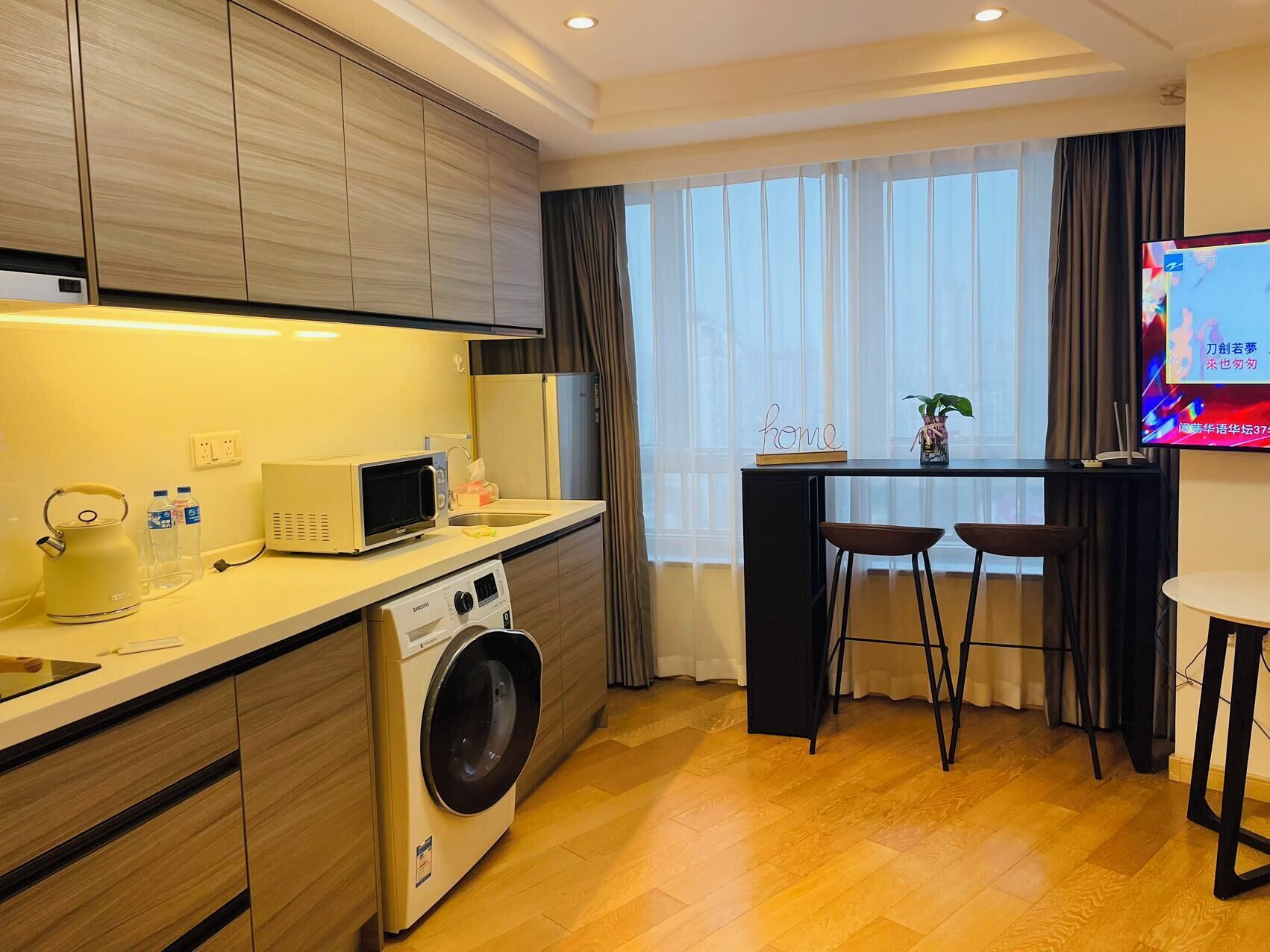 A compact kitchen with a microwave, kettle, and washing machine. There is a small bar area with stools and a TV.