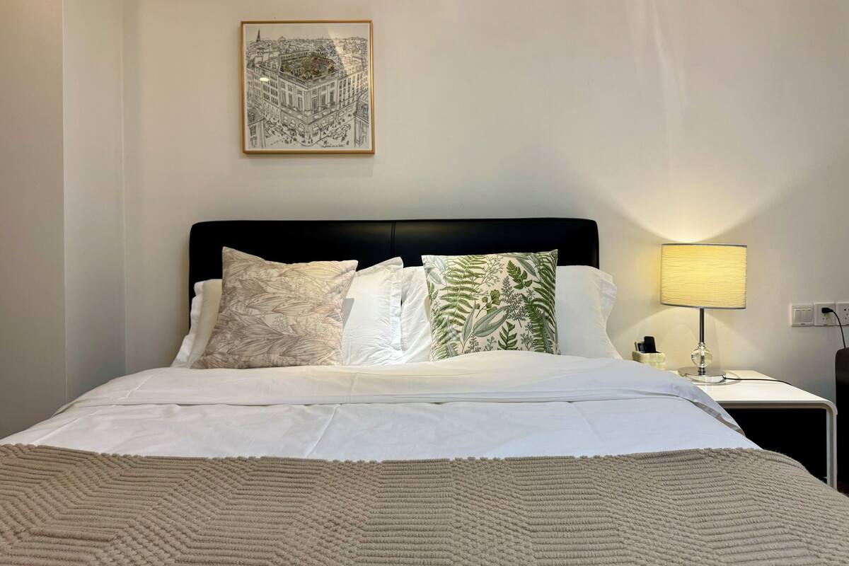 Un lit bien fait, avec du linge de lit blanc et un plaid beige à texture intéressante. On y trouve trois oreillers aux motifs différents, et une œuvre d’art encadrée est accrochée au-dessus du lit. Une lampe de chevet est allumée sur la droite.