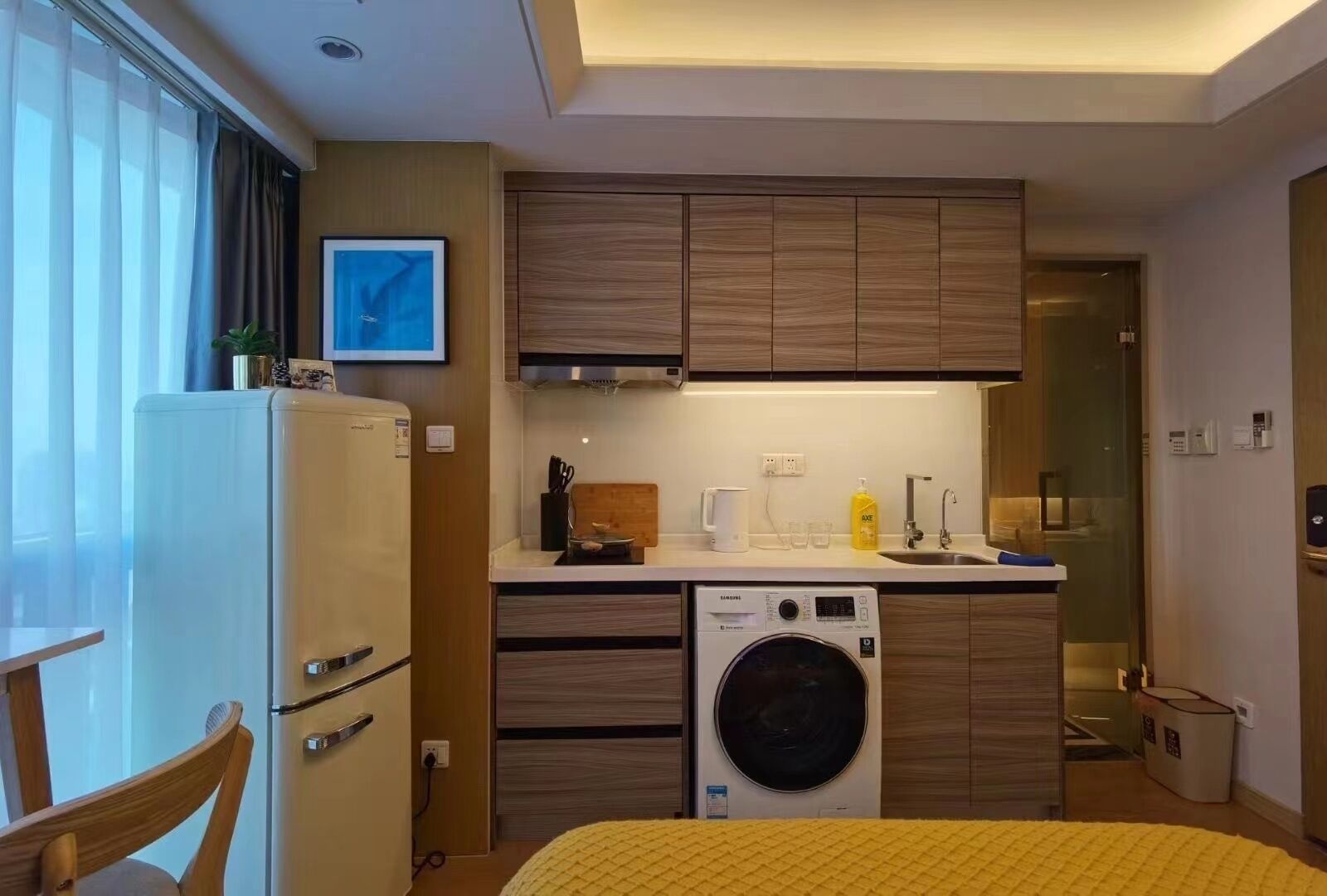 A compact hotel room with a kitchenette, a washing machine, a bed with a yellow blanket, and a dining area with a chair.