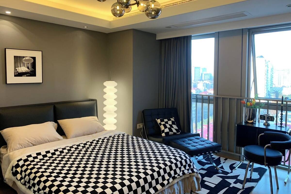 Une chambre d’hôtel moderne dotée d’un grand lit, d’un coin salon avec un fauteuil et un pouf, d’une vue sur la ville et d’un lustre original.