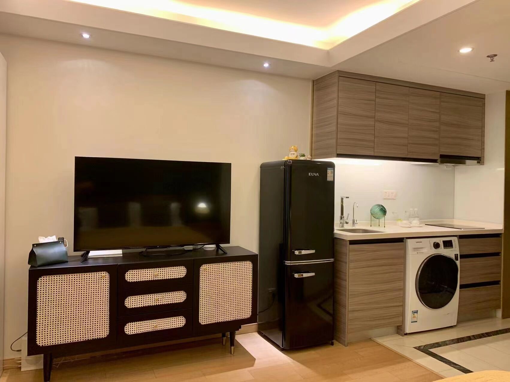 A modern kitchen with a built-in TV, a black refrigerator, a washing machine, and wooden cabinets.