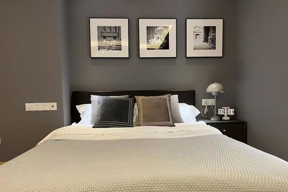 Un lit bien fait, avec une couette blanche à texture intéressante, une tête de lit sombre et plusieurs oreillers. Trois tableaux encadrés sont accrochés au mur au-dessus du lit.
