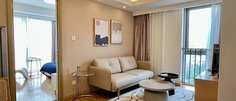 A modern living room with a sofa, a round coffee table, and a TV. There is a balcony with a view, and the room has a large window.