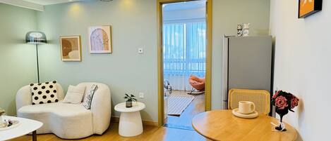 A modern living room with a sofa, a round wooden table, and a chair. There is a small white table with a plant, a floor lamp, and artwork on the walls.