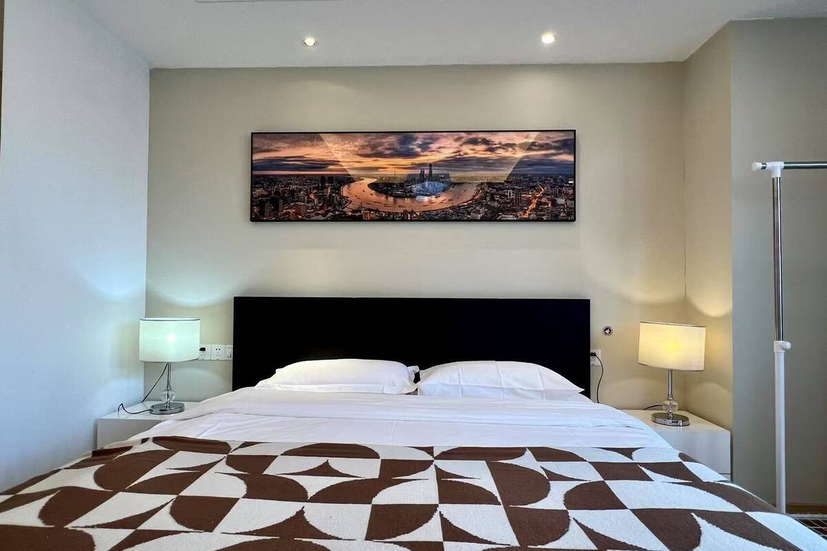 Une chambre d’hôtel avec un tableau représentant un paysage urbain, un grand lit recouvert d’une couvre-lit à motifs et deux lampes de chevet.