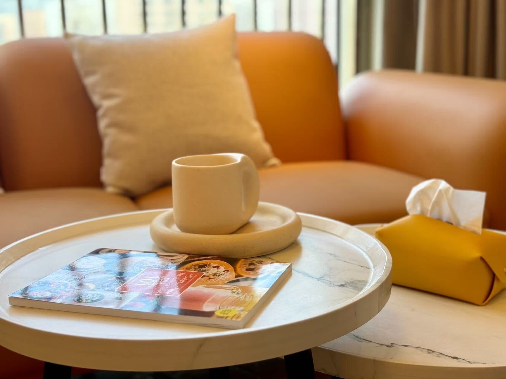 A round table with a magazine, a white mug, and a yellow tissue box.