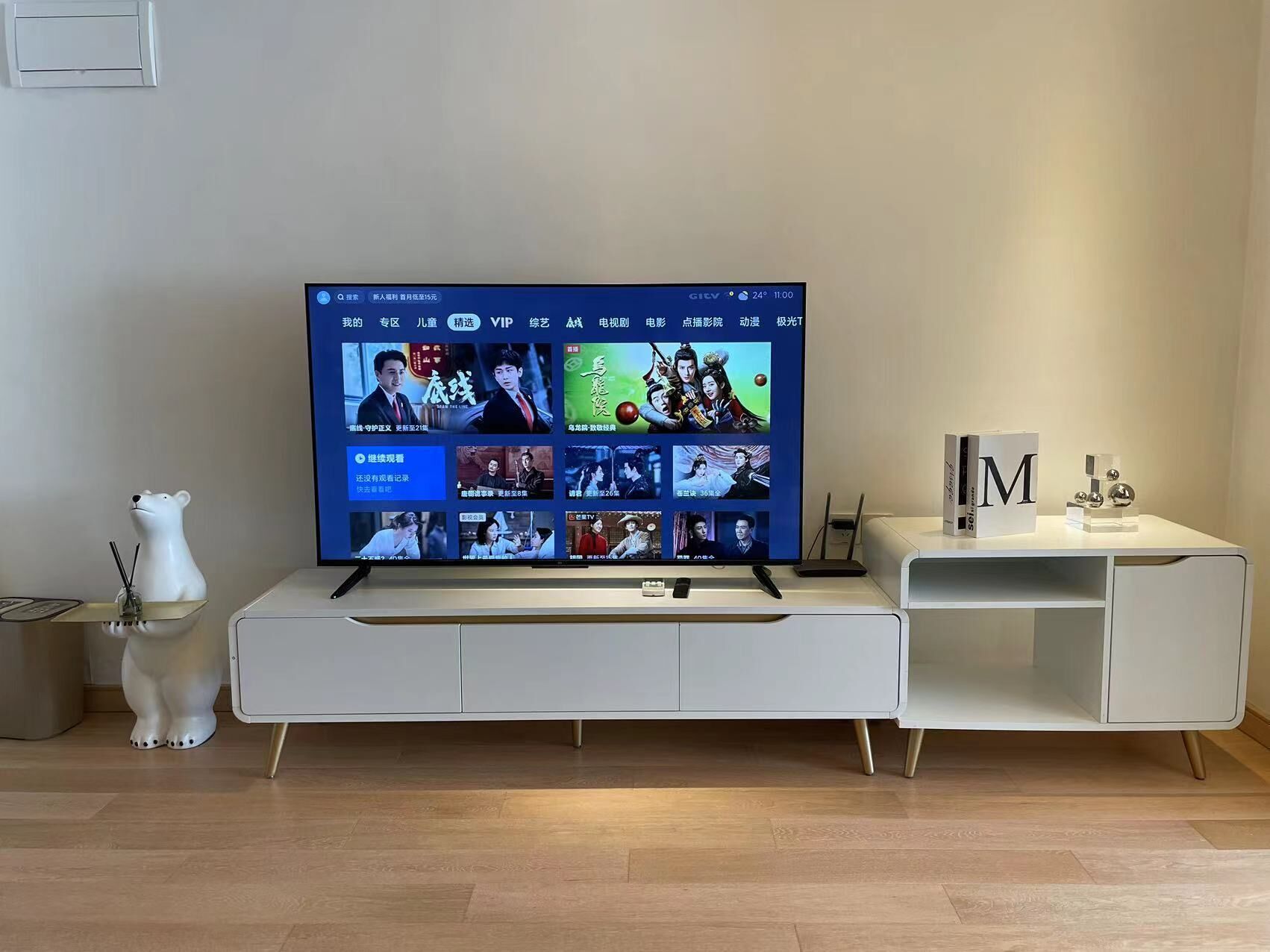 A flat-screen TV on a white entertainment center with a remote control and a decorative bear figurine on the left.