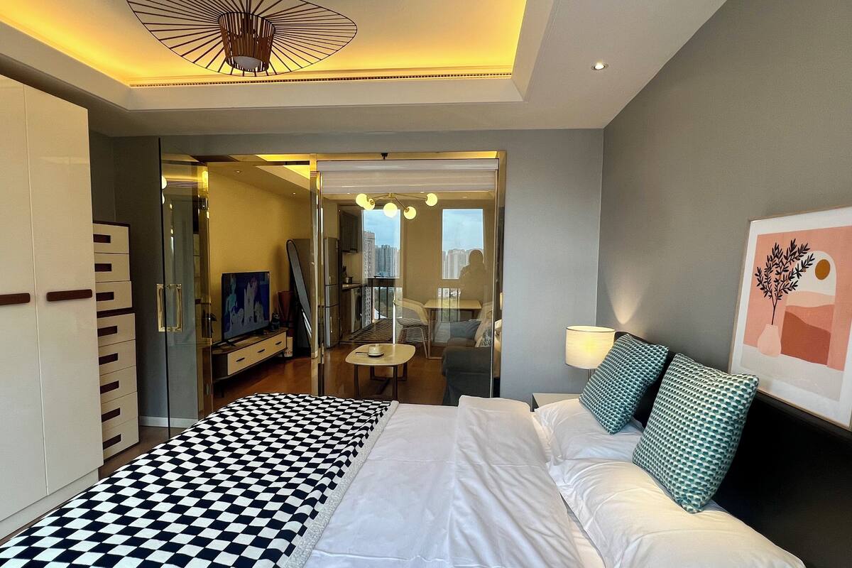 Une chambre d’hôtel moderne avec une couvre-lit à carreaux, un téléviseur à écran plat, une cloison en verre et une vue sur le paysage urbain.